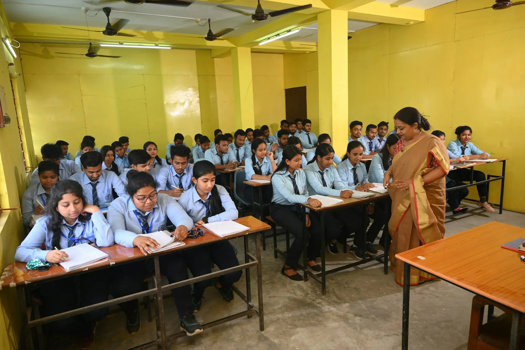 A teacher standing between rows of desks in a classroom with yellow walls