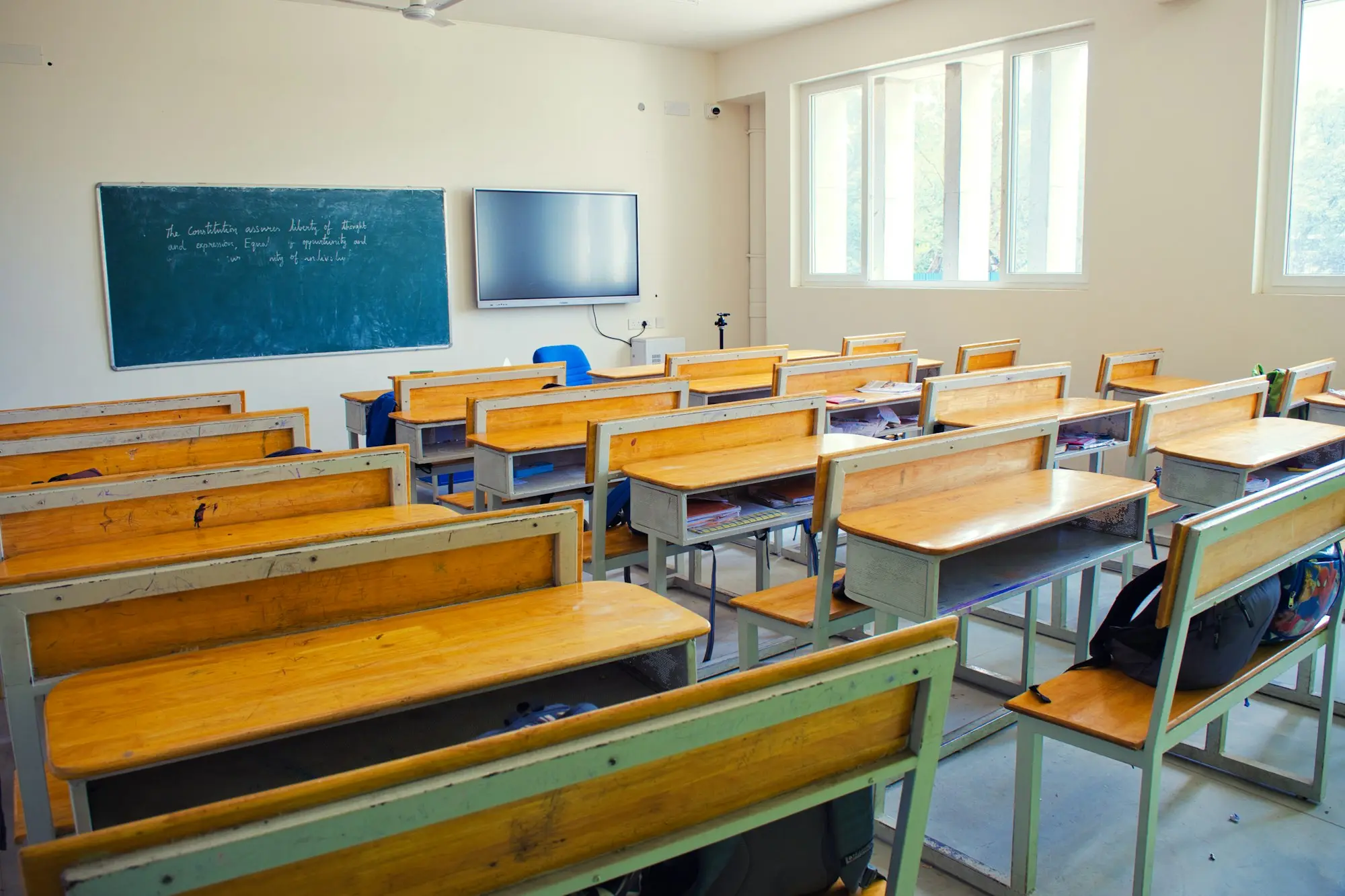 Rows of empty wooden desks in a classroom with daylight through the windows