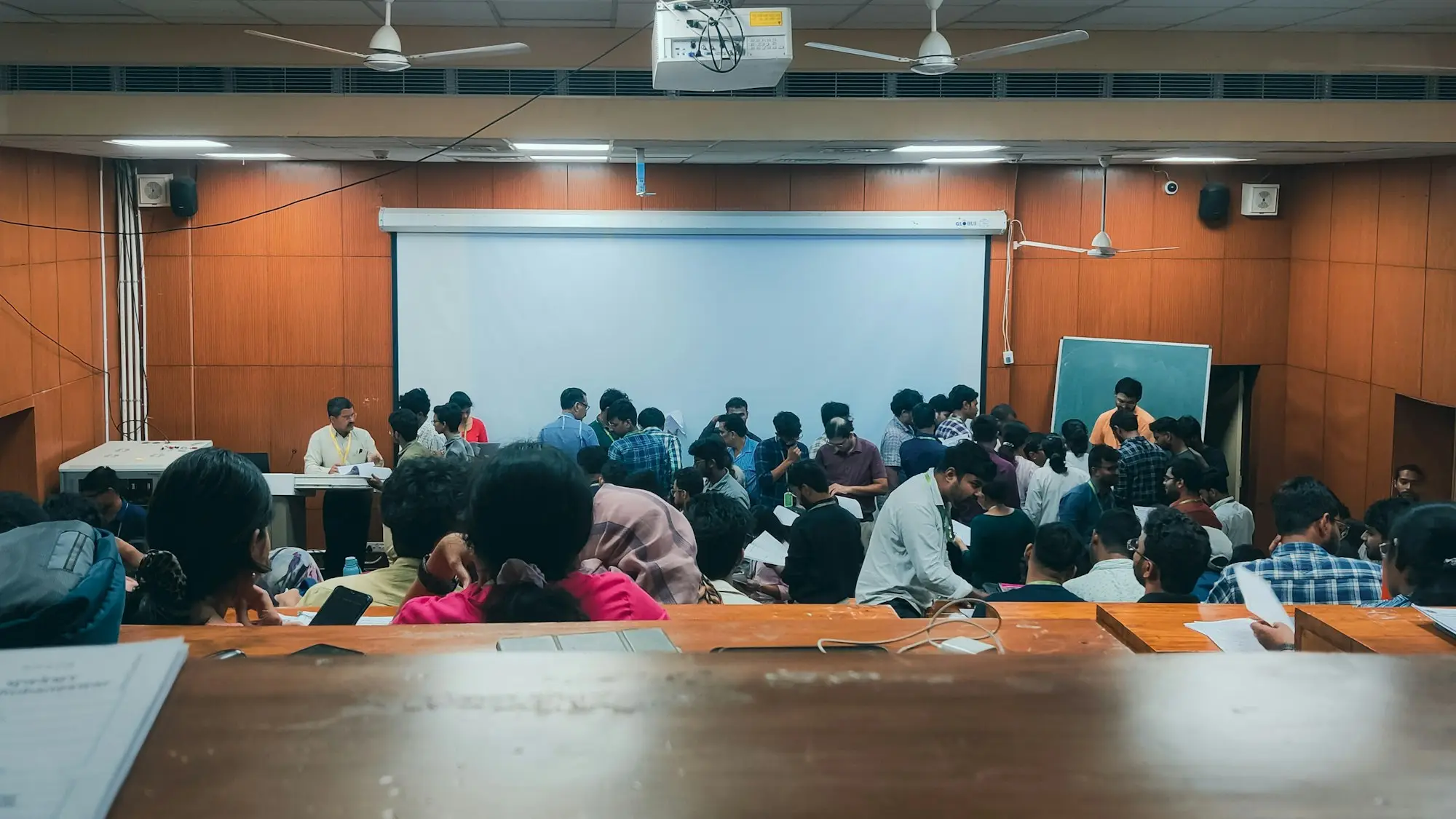 A room of people seated at long tables facing a projector screen